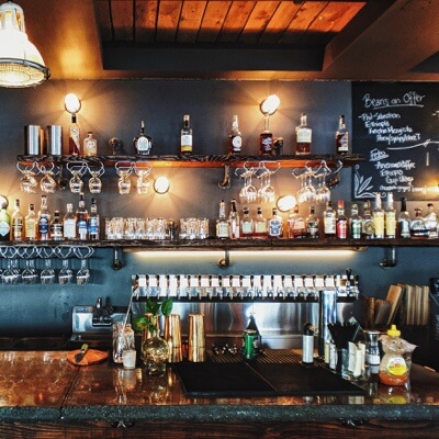 A shiny bar top with a prep station, backdropped by many different glasses and alcohol bottles on shelves