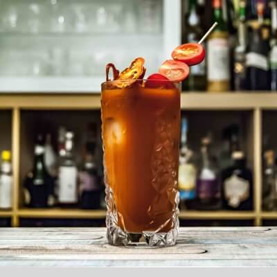 A dark red-orange drink in a tall glass, with a skewer of tomatoes placed in it, set on a bar top.