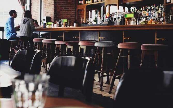 Two people sit on the far left side of a bar by a bright window, many empty bar stools are lined up to the right of them.